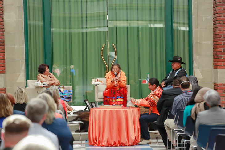Three speakers sit at the front of a room with an audience in the foreground