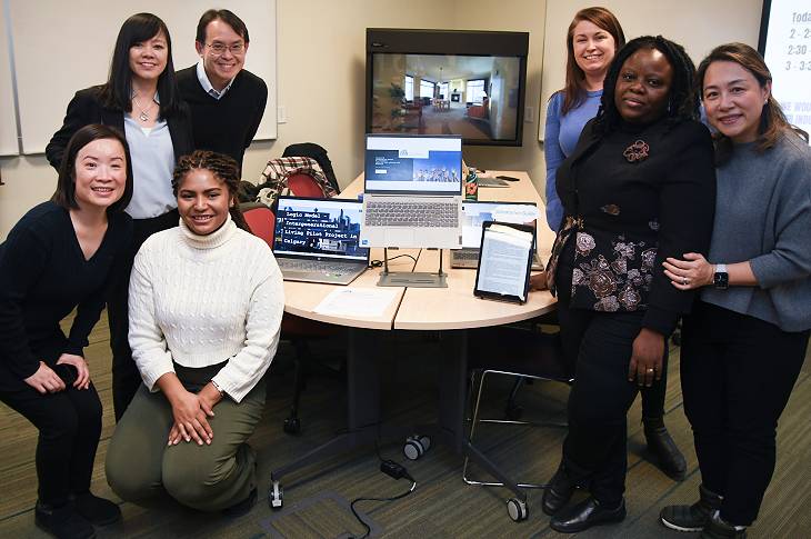 Seven students stand around a table. On the table, there are two laptops and a tablet; on their screens, report content (illegible).