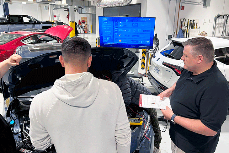 Honda technicians gathered around the front of a car with the hood up with a diagnostic screen in the background