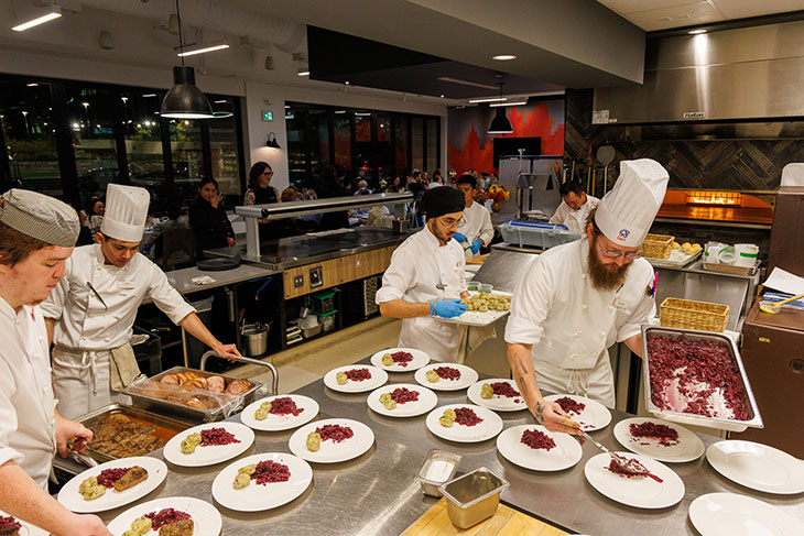 students in chef white and gloves plating a dish for an event with people seated in the background at The Tastemarket