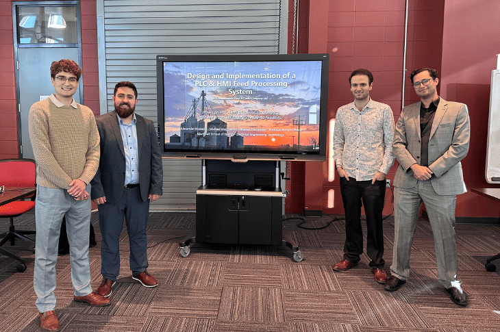 Four SAIT students stand in front of a red brick wall. They are surrounding a large television screen, which has a slide that says "Design and Implementation of a PLC & HDI Feed Processing System" on it.