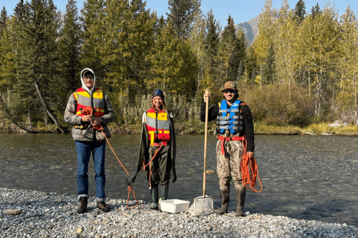 Three SAIT students stand on the rocky shore of a river, each wearing a life jacket and waterproof boots. Behind them, a mountain peeks through the trees. The day is bright and sunny.