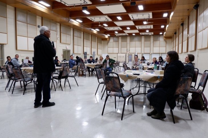 A speaker stands in front of a classroom full of learners, who are all seated at tables with markers and papers.