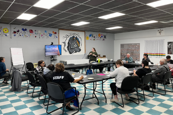 Group meeting in a classroom setting with individuals seated at tables, engaged in a presentation. A painted mural of a wolf and various artwork decorate the walls.