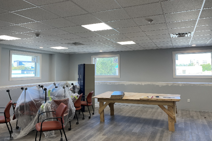 Photo of a classroom with three clear windows. Through the windows, green trees. Inside the classroom, chairs are wrapped in plastic and a workbench is in the centre.