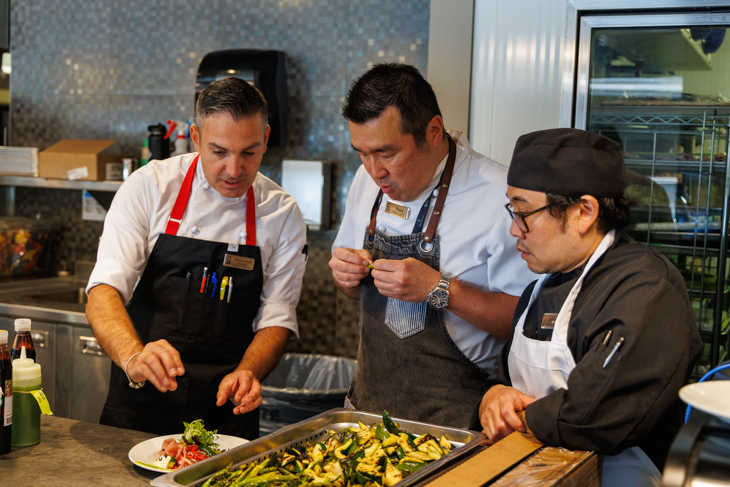 Three people prepare dishes at a counter in chef uniforms