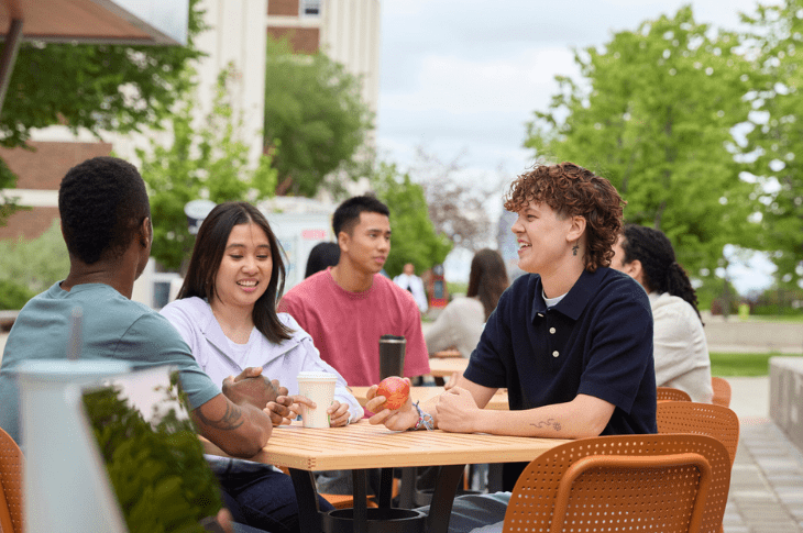 A group of students enjoying conversation and coffee outside on SAIT's campus. They are sitting at a table with green trees around them.