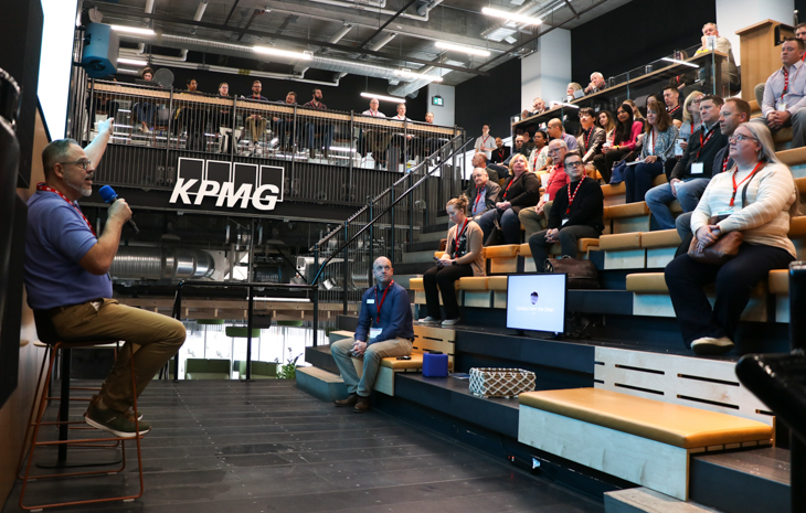 A speaker presents to an audience seated in a modern auditorium space, featuring an open layout with multiple levels of seating.