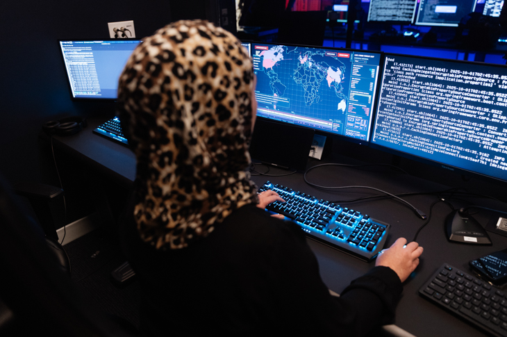 A woman wearing a leopard-print hijab sits at a desk with multiple computer screens displaying code and a world map.