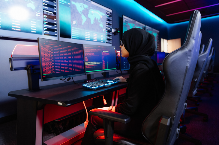 A woman in a hijab focused on multiple screens displaying data and maps in a high-tech control room.