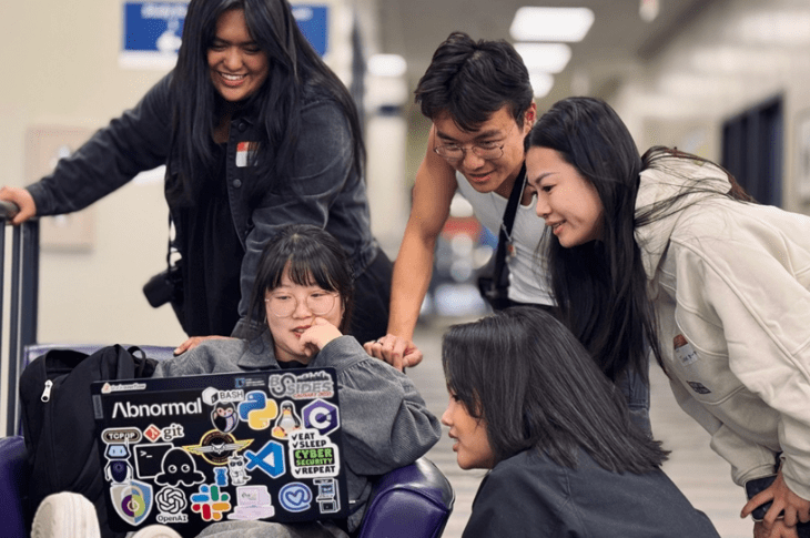 Five students gather around a laptop. They are all looking at something on screen and smiling.