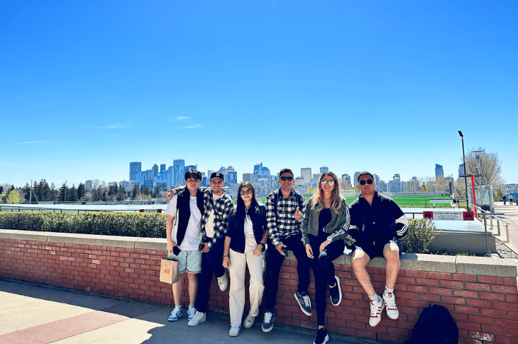 A group of six students lean against a brick wall on SAIT's Main Campus in front of blue skies and a view of the downtown Calgary skyline. 