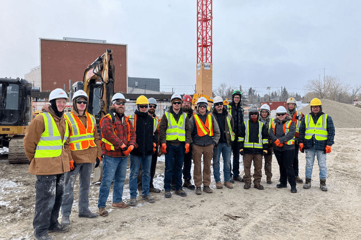 A group of 15 SAIT students and employees stand on a construction site in the wintertime. They are all wearing hard hats, fluorescent vests and steel-toed boots. Behind them, there is a crane and a backhoe. 