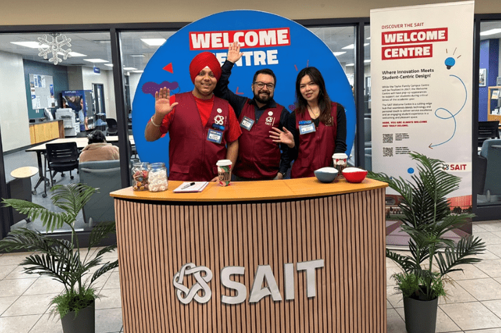 Three SAIT employees stand behind a wooden booth that says "SAIT." Behind them, there is a blue sign that reads "Welcome Centre." Another sign in the background repeats "Welcome Centre." There are potted plants on either side of the group.
