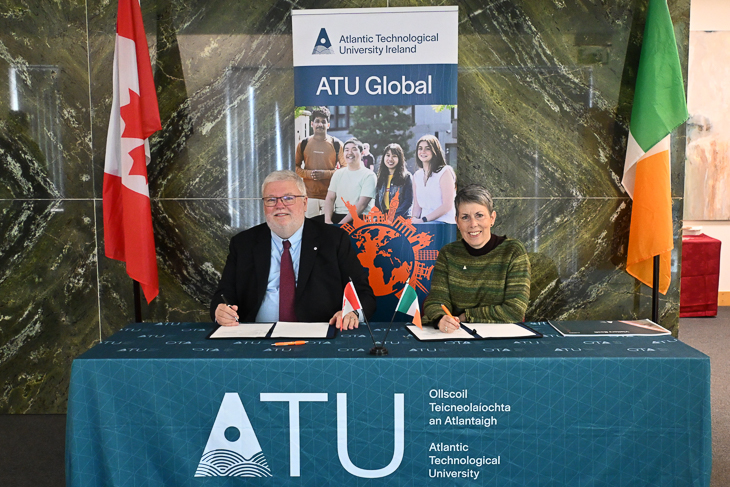 Two officials sign documents at table with a banner for Atlantic Technological University Ireland and Canadian and Irish flags behind them.