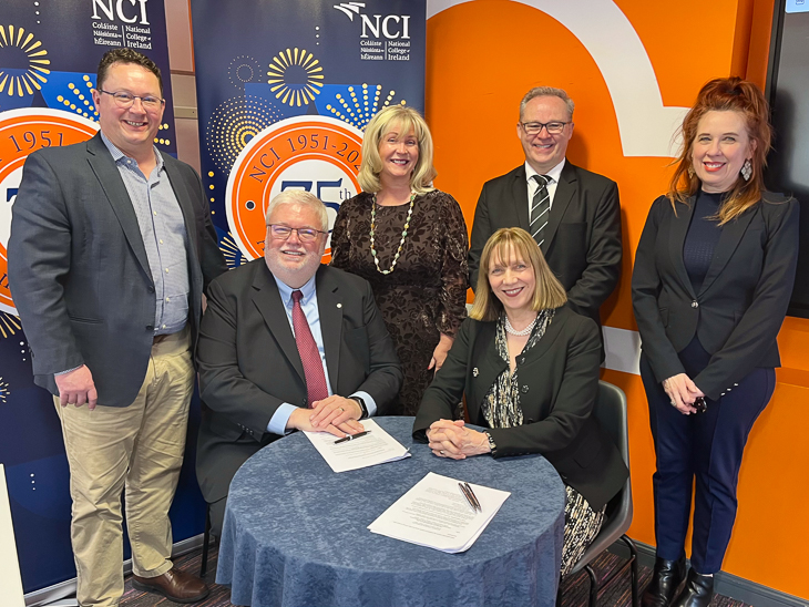 Group photo with two people seated at a table signing documents and four colleagues standing behind them.