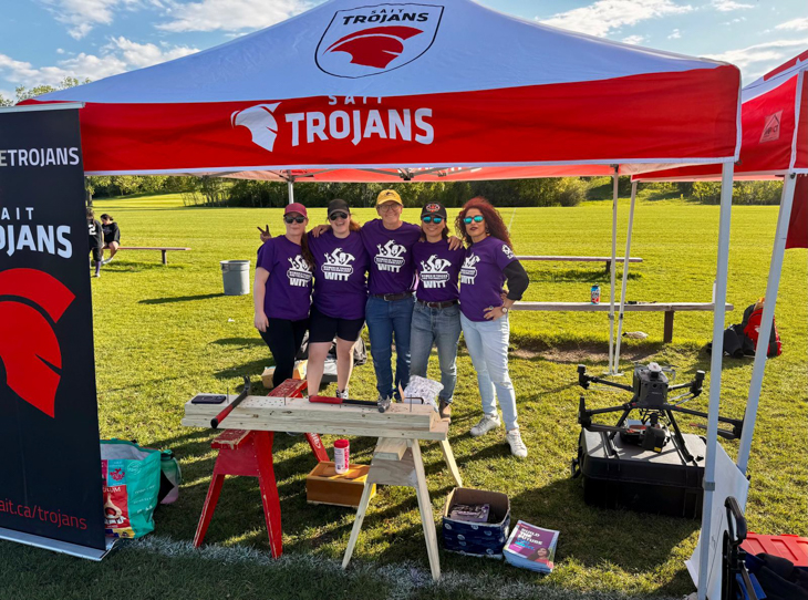 A group pose for a photo outdoors at a SAIT Trojans game wearing women in trades and technology shirts