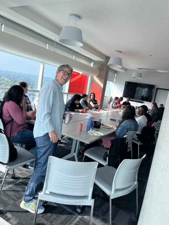 A person is standing up smiling at the camera with a group meeting around a boardroom table behind them