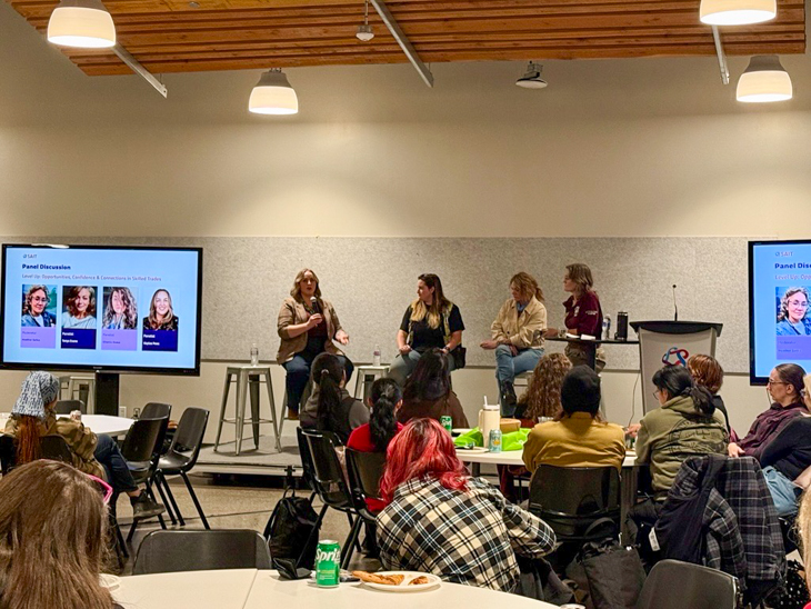 People sitting a round tables in a meeting space listening to a panel talk about women in skilled trades careers