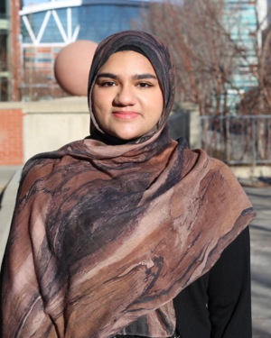 Yumnaa Farooq stands outside of SAIT's Aldred Centre on a sunny day.