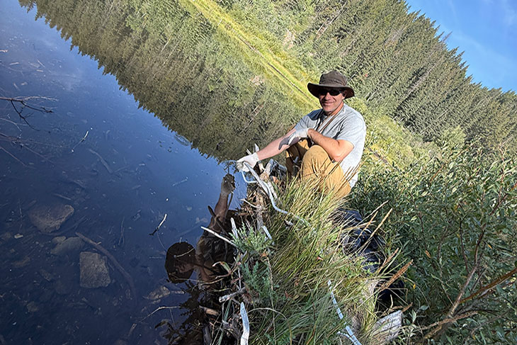 Colin Pattison by water with trees and blue sky in the background.