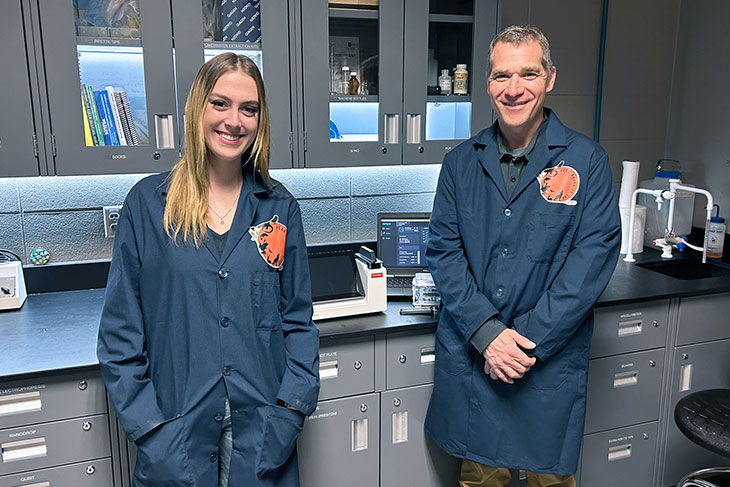 Olivia Zamrykut (left) and Colin Pattison (right) wearing navy blue lab coats in SAIT's eDNA lab. The background features gray cabinets, scientific equipment, and a computer monitor displaying data, indicating a research or scientific work environment.