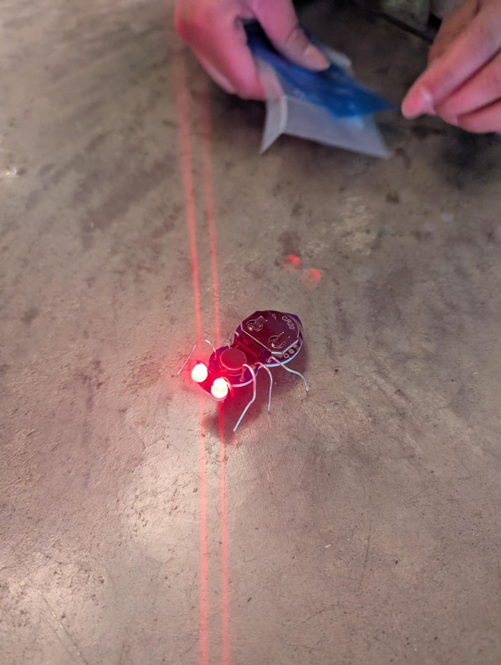 An electronic insect sits on the floor with eyes lit up in red in an avionics lab.