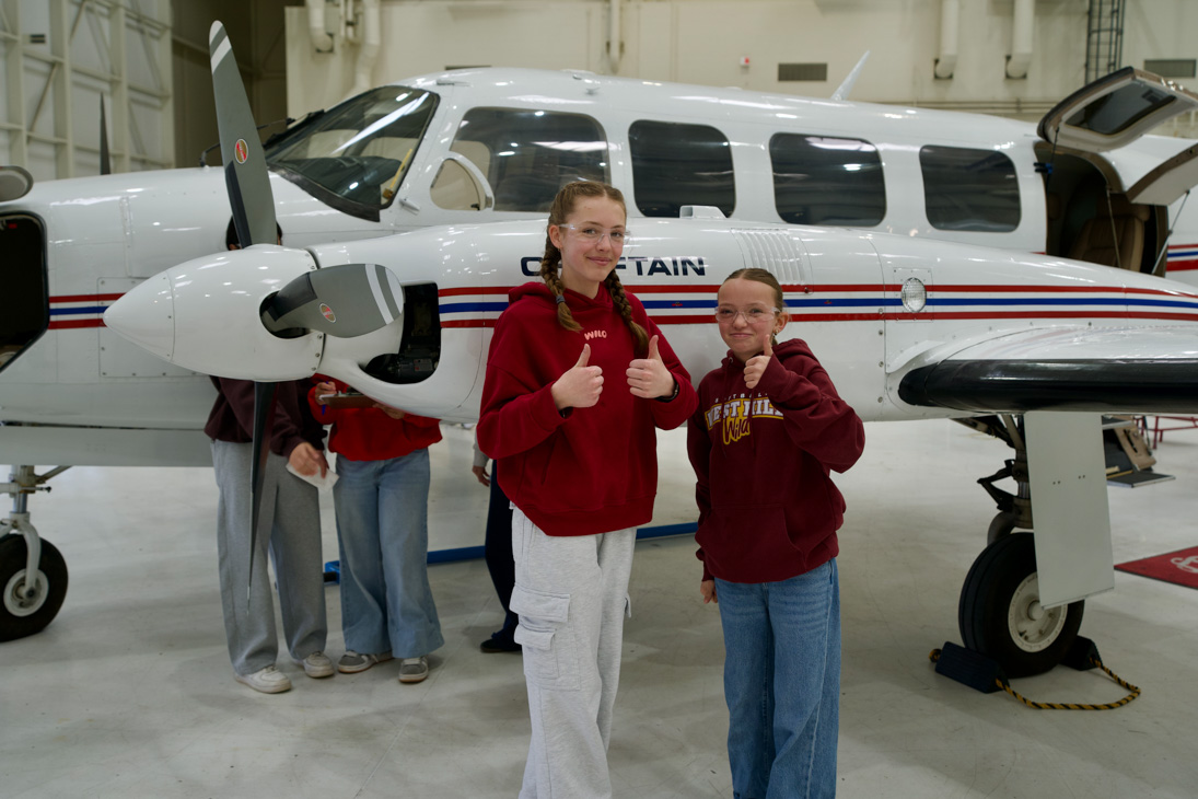 Students give a thumbs up standing in front of a plane.