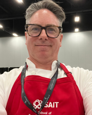 A selfie of Patrick Creery in SAIT's kitchens. He is wearing a red apron with SAIT embroidered on the centre.