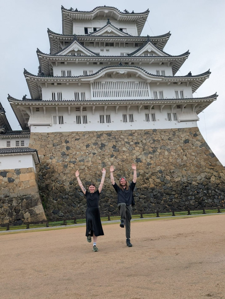 Two people posing with raised arms in front of a large white Japanese castle.