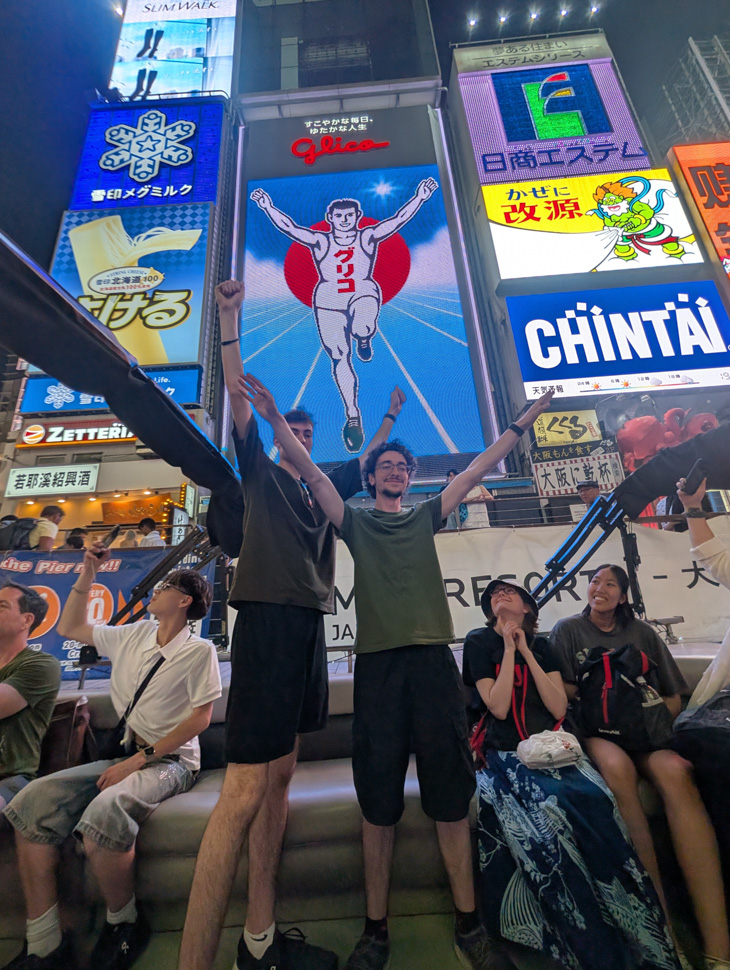 Group of people posing together beneath colourful illuminated signs on a busy city street.