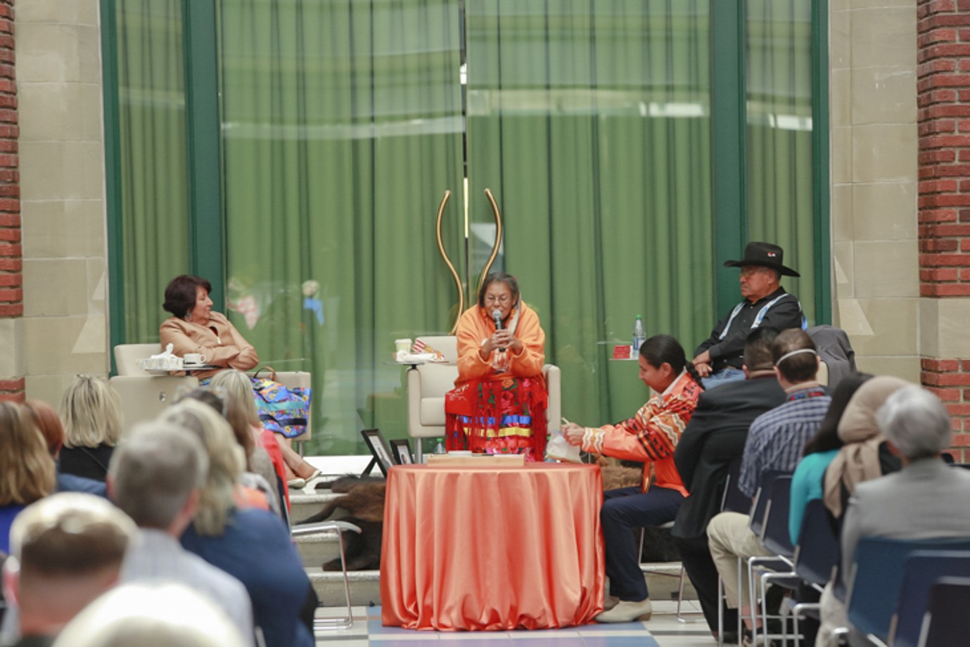SAIT's resident elder, Betty Lou Crazyboy, sits in front of an orange table, speaking into a microphone. 