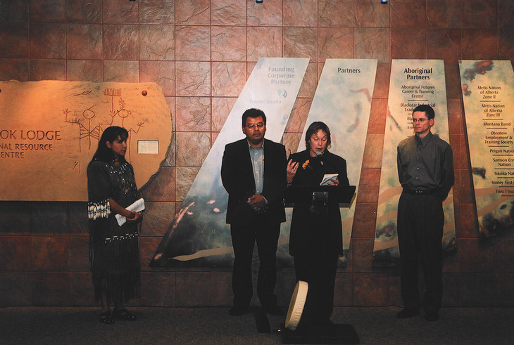 Three people stand together as one speaks into a podium at the grand opening of SAIT's Indigenous student support centre