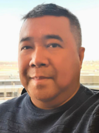 A smiling man with short hair, wearing a navy blue sweater, poses for a selfie against a bright backdrop.
