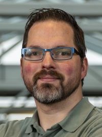 A man with glasses and a beard smiles confidently in a professional setting.