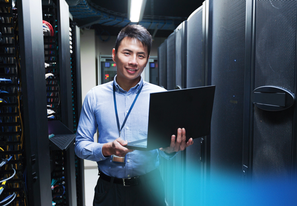 A smiling IT professional holds a laptop while standing in a server room filled with electronic equipment.