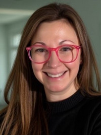 A smiling woman wearing large pink glasses and a black sweater, with a soft background.