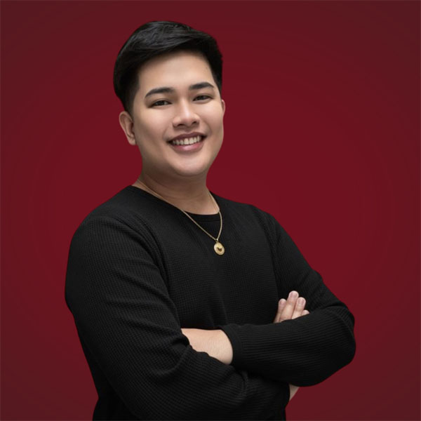 Al Kim Lutao with their arms crossed, wearing a black sweater and a gold necklace against a red background.