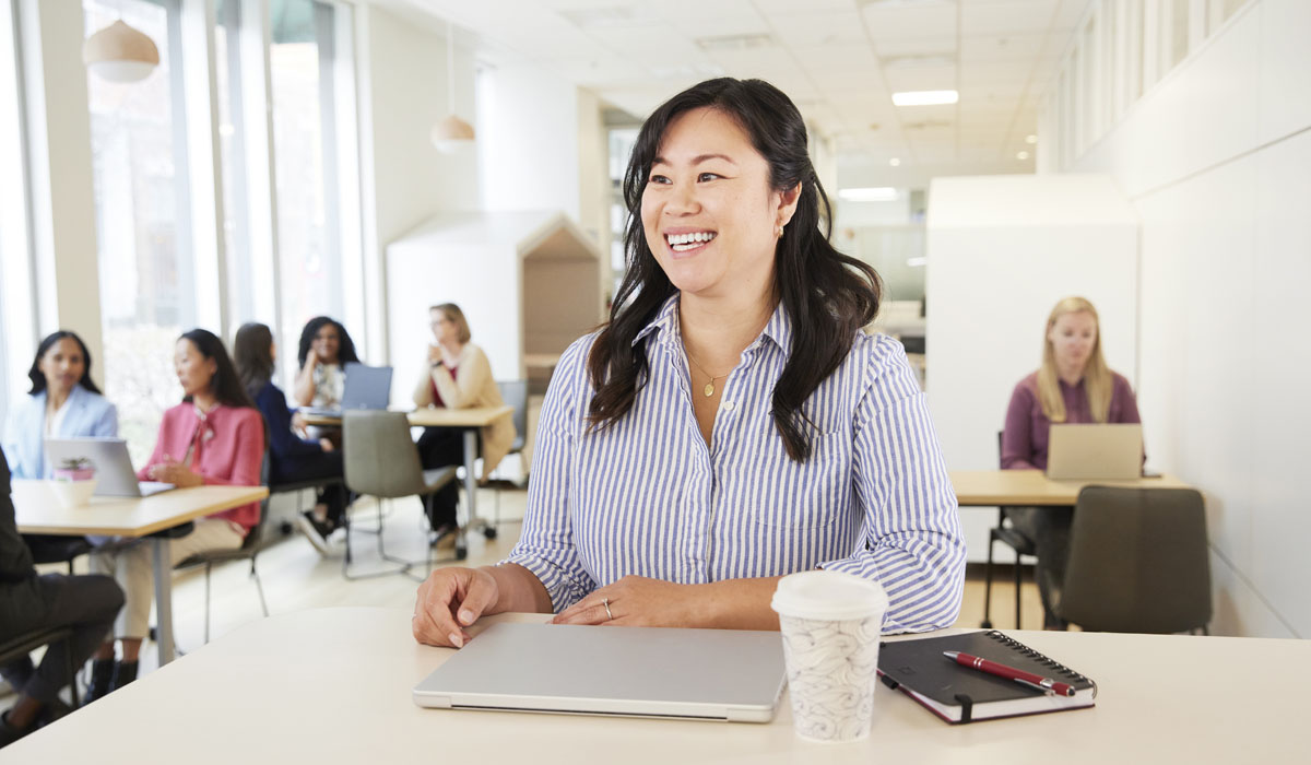 A smiling woman at a table with a laptop, coffee cup, and notebook, surrounded by people working in a bright, modern office space.