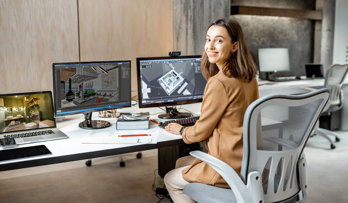A professional woman sitting at a modern workstation with multiple monitors displaying design software and a laptop nearby.