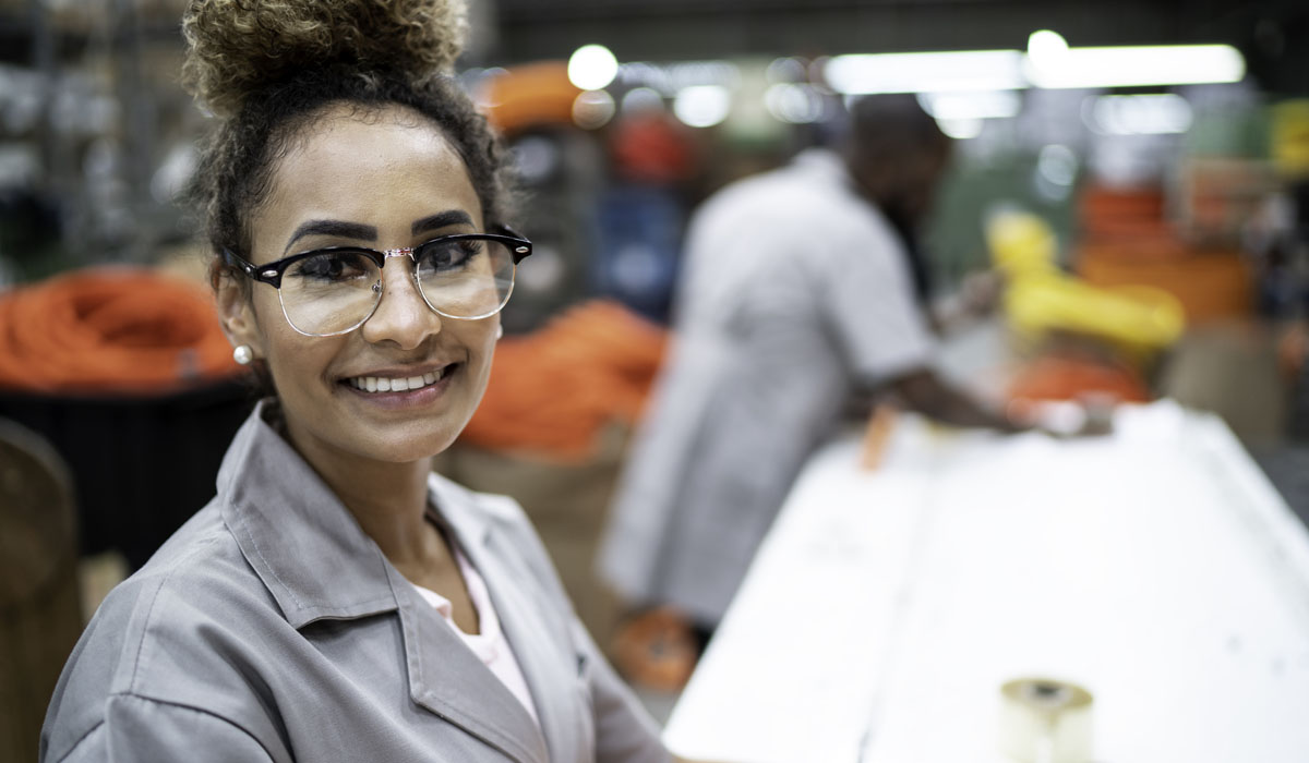 A smiling woman wearing glasses and a work uniform in a manufacturing setting. Background features another worker and colourful materials.