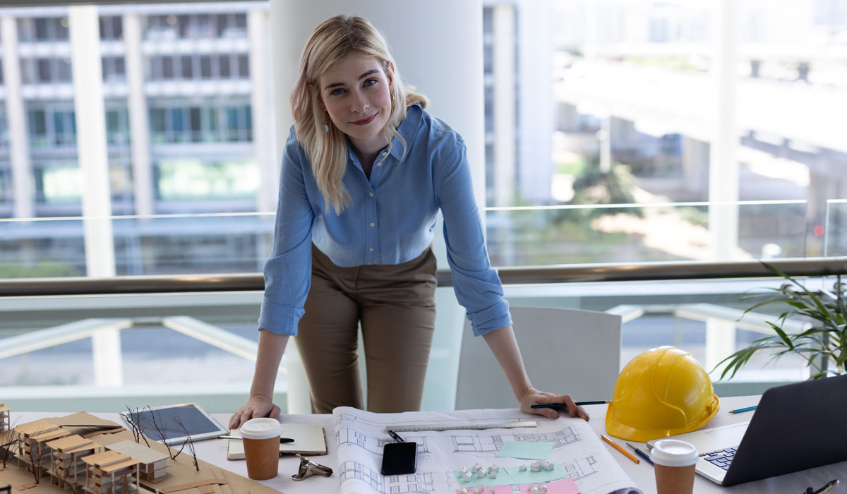 A woman in a blue blouse leans over architectural plans on a table, with a hard hat and coffee cups nearby, in a modern office setting.