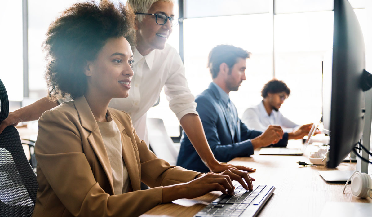 A diverse group of professionals collaborating in a modern office, with a focus on a woman working on a computer while a colleague provides guidance.