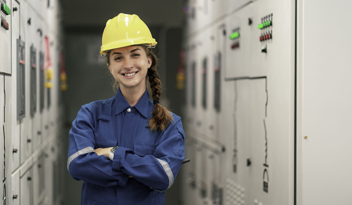 A smiling female electrician wearing a hard hat and blue work uniform, standing confidently in a corridor lined with electrical panels.