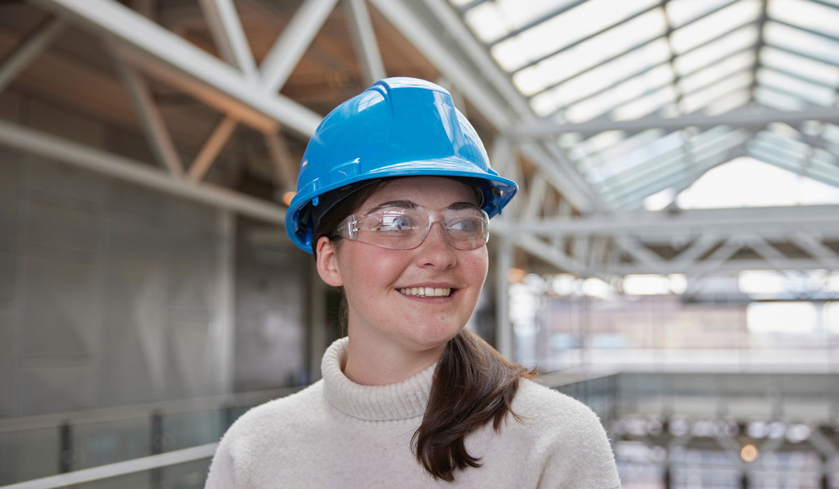 A smiling woman wearing a blue hard hat and safety glasses in a modern building environment.