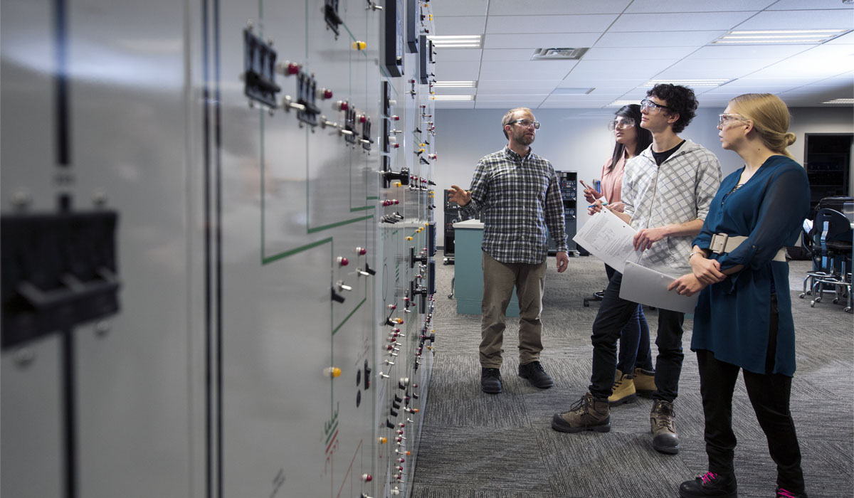 A group of four individuals engaged in a discussion in a lab setting, examining a control panel with various switches and indicators.