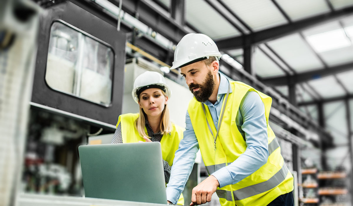 Two student workers wearing safety helmets and vests collaborate over a laptop in an industrial setting.