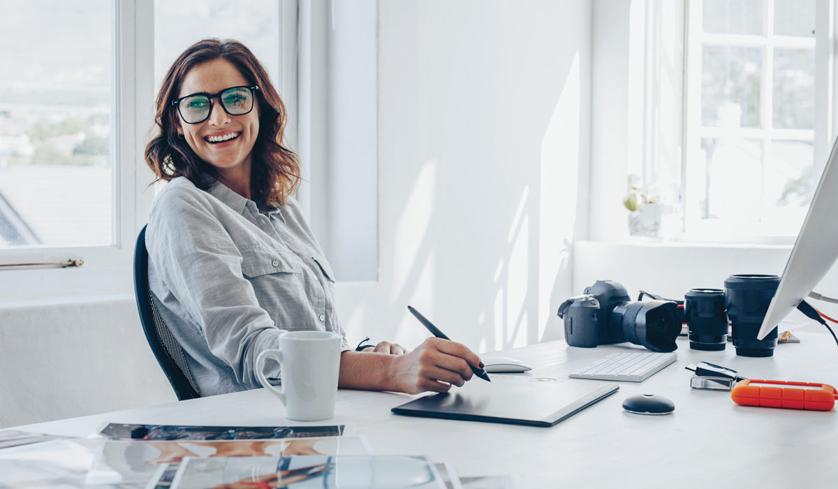 A smiling woman with glasses is sitting at a desk, working on a digital tablet with photography equipment nearby.