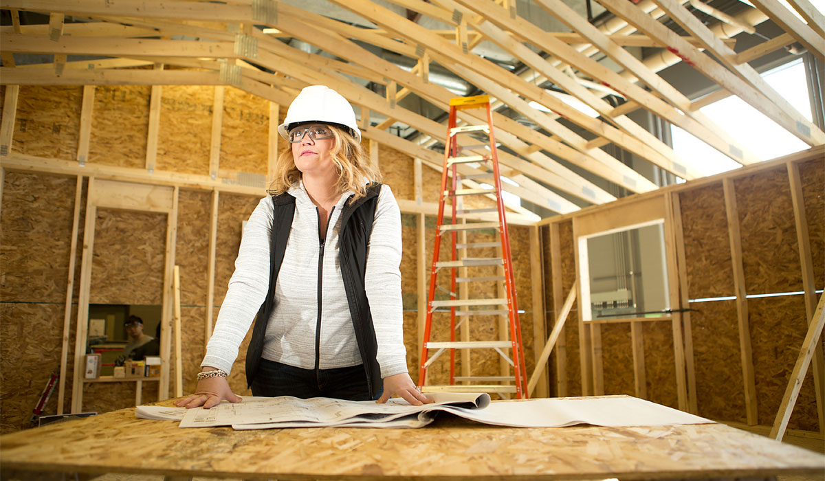 A woman in a hard hat stands at a construction site, looking over blueprints on a wooden table, with a ladder and wooden framing in the background.