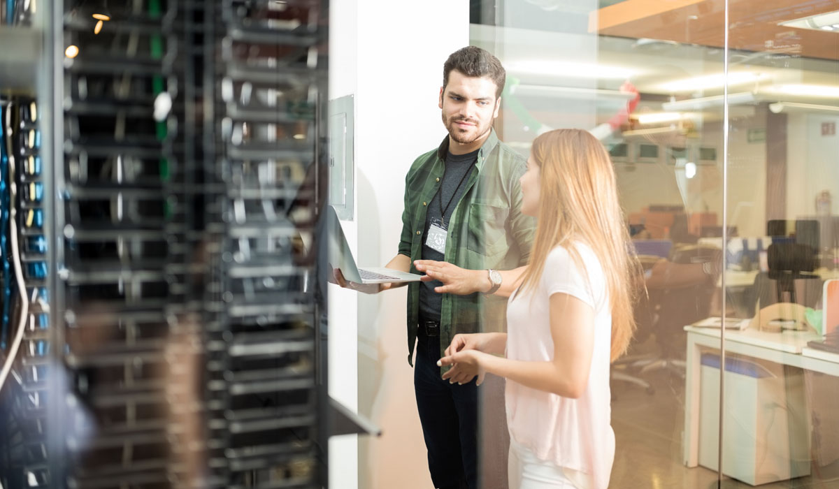 Two professionals discussing computer systems in a data center, with server racks in the background.